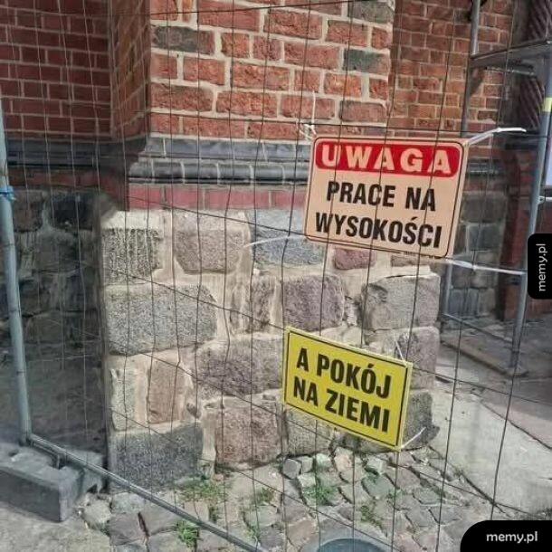 Dwie tabliczki na ogrodzeniu budowlanym przy ceglanym budynku.