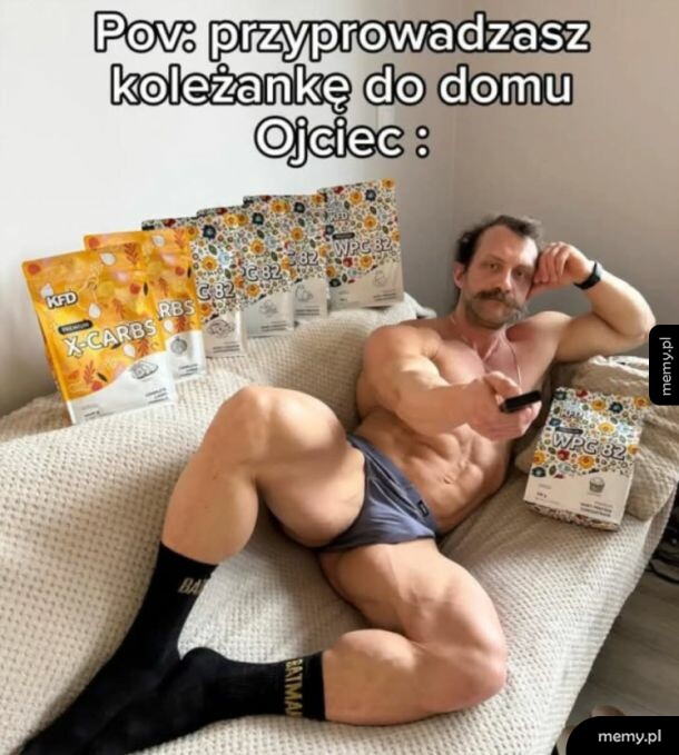 Mężczyzna w bieliźnie siedzi na kanapie z odżywkami białkowymi w tle.